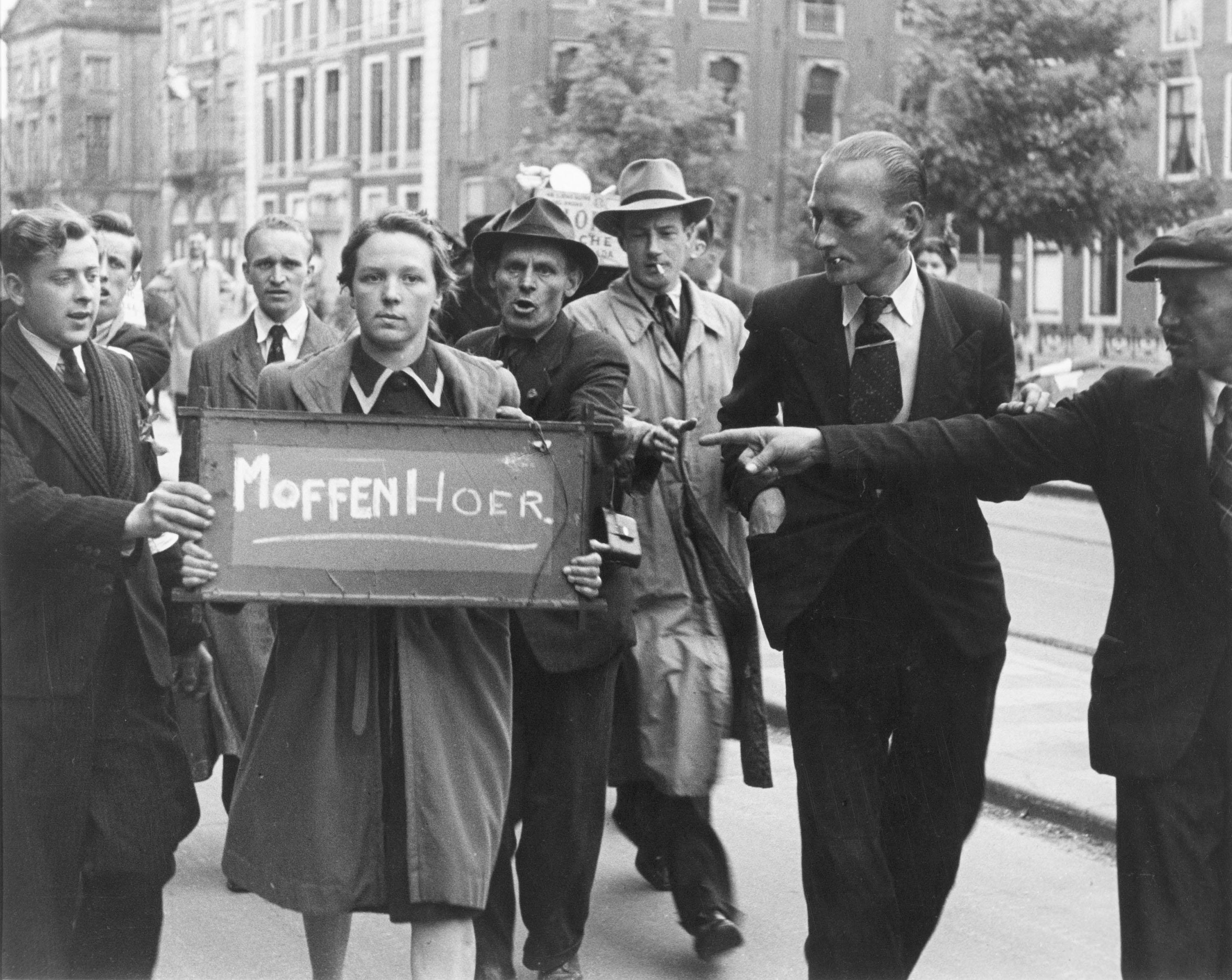 een vrouw draagt een bord met 'moffenhoer', mannen staan eromheen en wijzen ernaar