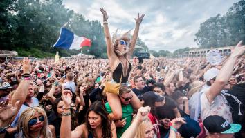 festivalgangers aan het dansen bij een optreden van een dj op een dancefestival. Een vrouw zit op iemands nek met haar handen juichend in de lucht.