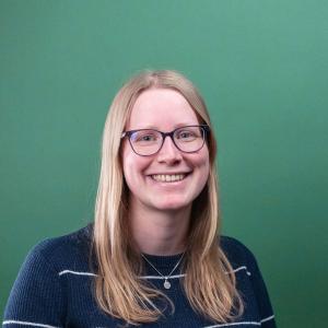 een portretfoto van een witte vrouw met blond haar en een bril met een donkergroene achtergrond