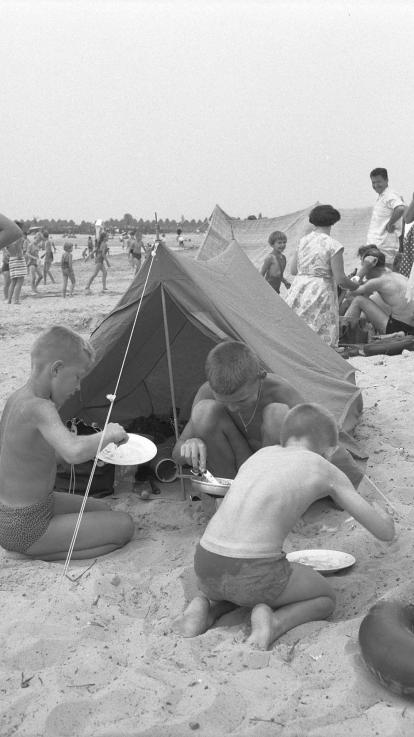 Drie jongetjes eten van hun borden voor hun kampeertent op het strand, op de achtergrond meer vakantiegangers