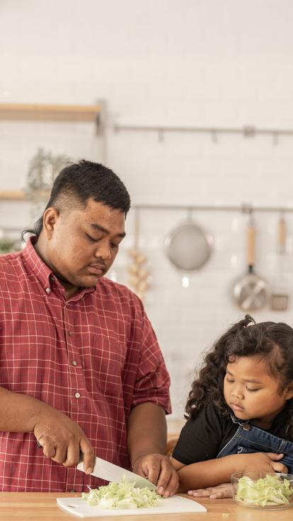 Een vader met overgewicht snijdt groente in de keuken, met zijn dochter naast hem