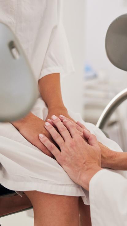 Vrouw zit op gynaecologenstoel en oudere vrouwenhanden houden haar handen vast