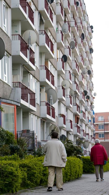 een appartementencomplex met balkons met schotels erop. Twee mensen lopen op straat voorbij.
