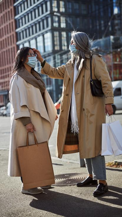 Twee vrouwen lopen met medisch mond-neusmasker op straat. De ene vrouw veegt een pluk haar achter het oor van de ander en ze hebben shoptassen bij zich