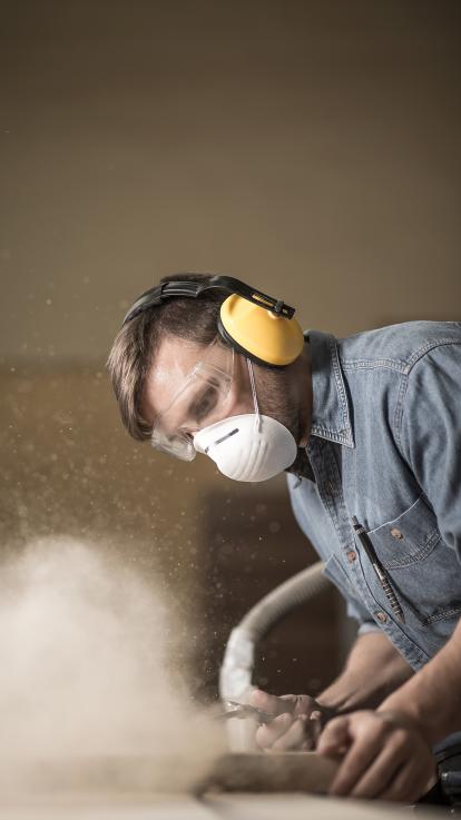 Een man schuurt een tafel met een beschermende koptelefoon, bril en stof-mondkap op. 
