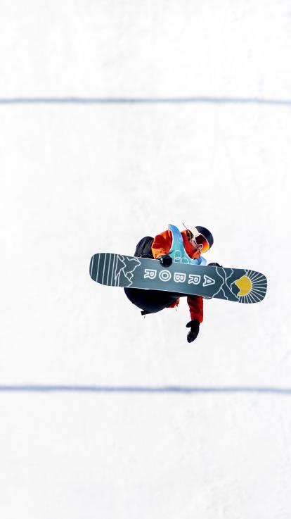 Een snowboarder maakt sprong in de lucht. Het board wijst naar de camera toe. 