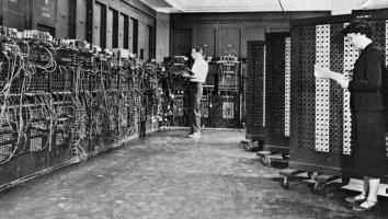 een zwart-witfoto van twee medewerkers die werken aan een van de eerste algemene elektronische computers, die bestaat uit allemaal grote kasten met kabels