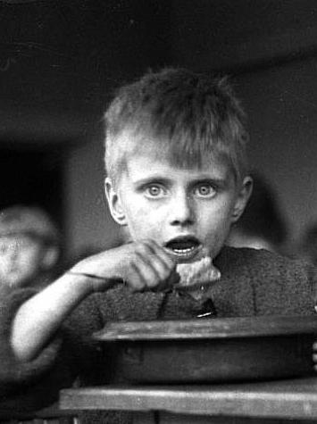een jongetje neemt een hap van zijn eten terwijl hij met grote ogen naar de camera kijkt, zwart-witfoto