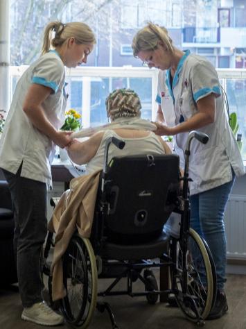 Twee verpleegkundigen helpen een vrouw in een roelstoel met aankleden