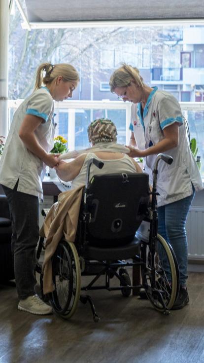 Twee verpleegkundigen helpen een vrouw in een roelstoel met aankleden