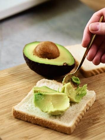 iemand legt stukjes avocado op zijn witte boterham, daarnaast ligt de andere helft van de avocado