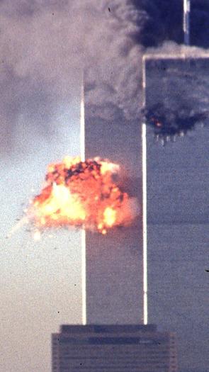 Een foto van de Twin Towers in 2001, direct na de tweede aanslag. Midden op de linkertoren is een vuurexplosie te zien. De rechtertoren staat in brand met dikke zwarte rookwolken.