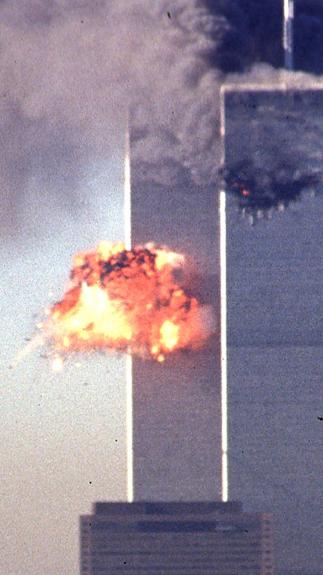 Een foto van de Twin Towers in 2001, direct na de tweede aanslag. Midden op de linkertoren is een vuurexplosie te zien. De rechtertoren staat in brand met dikke zwarte rookwolken.