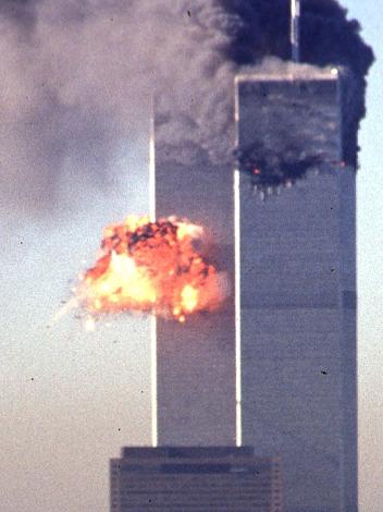 Een foto van de Twin Towers in 2001, direct na de tweede aanslag. Midden op de linkertoren is een vuurexplosie te zien. De rechtertoren staat in brand met dikke zwarte rookwolken.
