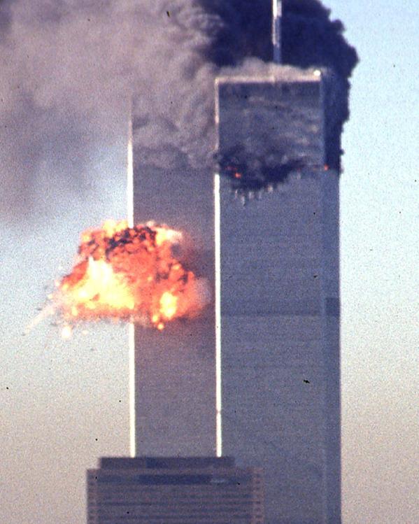 Een foto van de Twin Towers in 2001, direct na de tweede aanslag. Midden op de linkertoren is een vuurexplosie te zien. De rechtertoren staat in brand met dikke zwarte rookwolken.