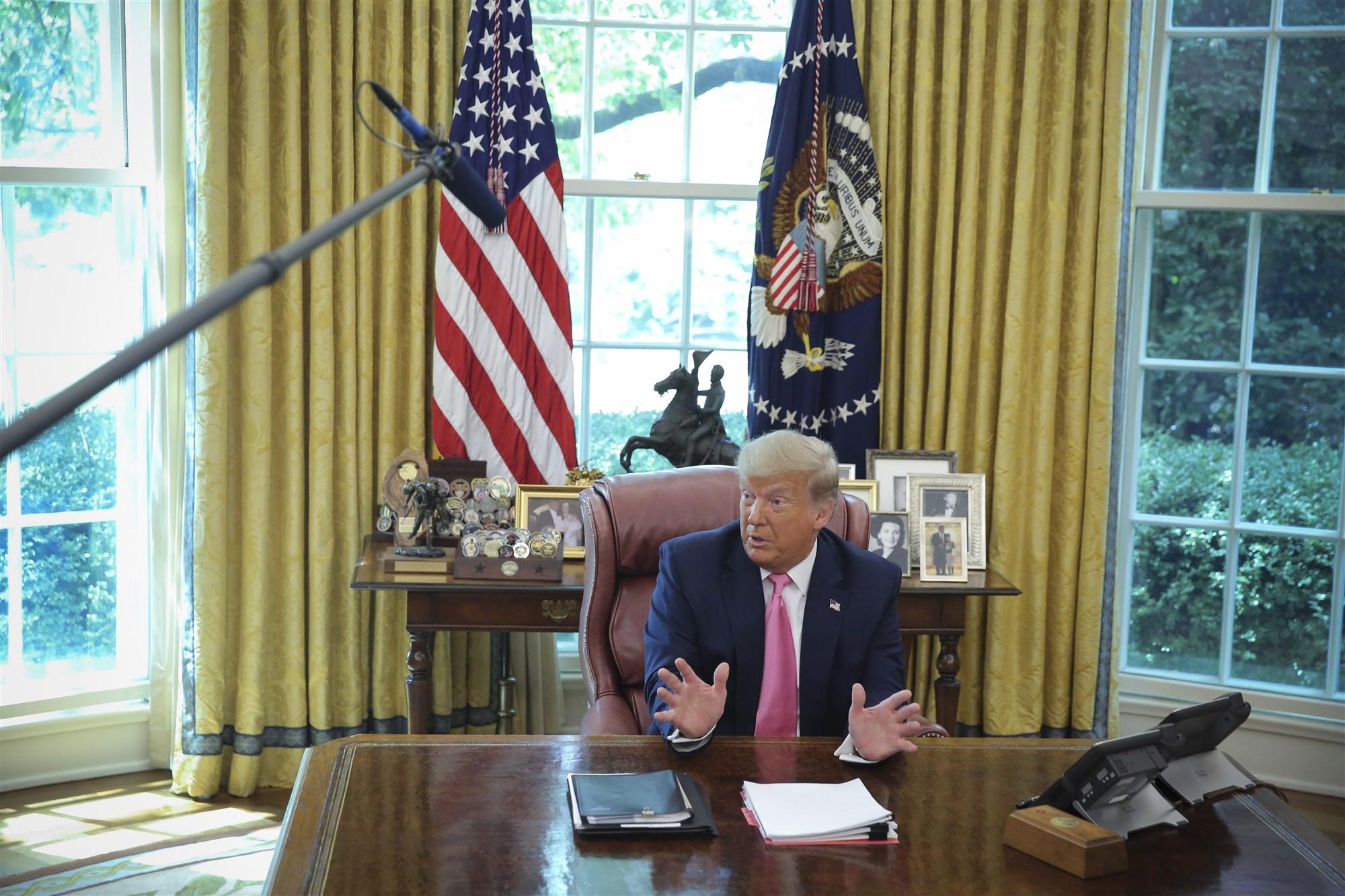 Een foto van president Trump die achter zijn bureau zit in de Oval Office en iets vertelt.