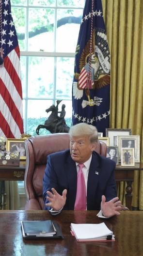 Een foto van president Trump die achter zijn bureau zit in de Oval Office en iets vertelt.