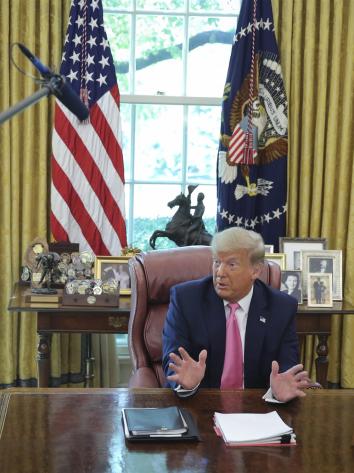 Een foto van president Trump die achter zijn bureau zit in de Oval Office en iets vertelt.