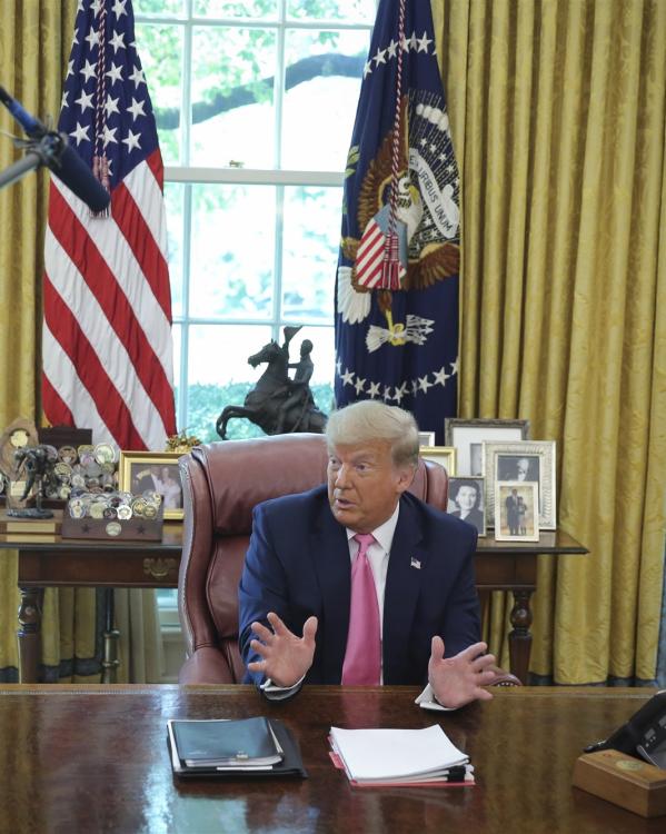 Een foto van president Trump die achter zijn bureau zit in de Oval Office en iets vertelt.