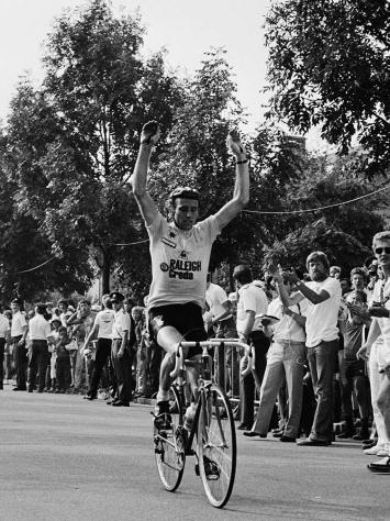 Joop Zoetemelk rijdt met zijn handen omhoog over de finish van de Tour de France. Er staat een hoop publiek en mannen op de tribune noteren de tijd. De foto is in zwart-wit.
