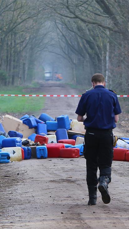 Er loopt een agent richting een hoop vaten drugsafval op een bospad