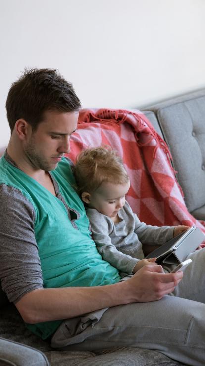 Vader zit op zijn telefoon en baby kijkt op tablet zittend op de bank