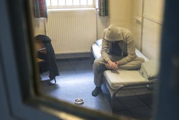 Door een gevangenisdeur zie je een man gebogen op bed zitten in een cel