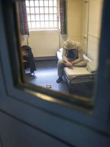 Door een gevangenisdeur zie je een man gebogen op bed zitten in een cel