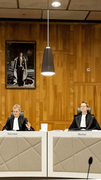 Twee rechters zitten aan hun tafel in een meervoudige rechtszaal, boven hen hangt een portret van de koning