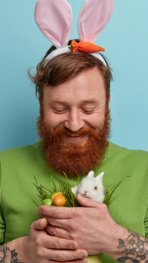 Man met baard heeft paasoren op en een konijntje en eitjes in zijn handen.