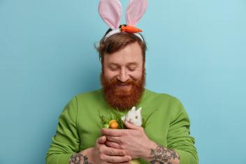 Man met baard heeft paasoren op en een konijntje en eitjes in zijn handen.