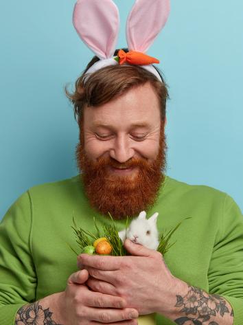 Man met baard heeft paasoren op en een konijntje en eitjes in zijn handen.