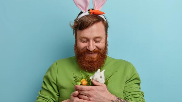 Man met baard heeft paasoren op en een konijntje en eitjes in zijn handen.