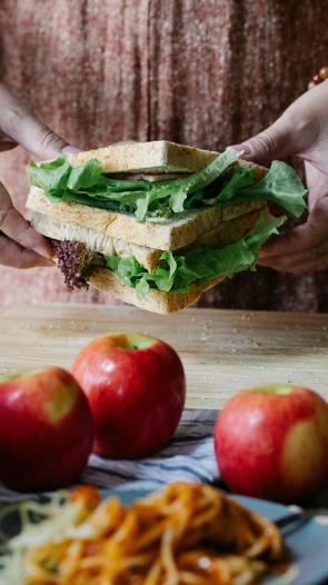 een tafel met producten waarachter je handen ziet die een sandwich vastpakken
