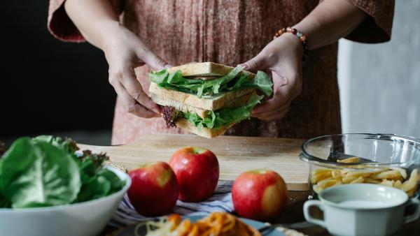 een tafel met producten waarachter je handen ziet die een sandwich vastpakken
