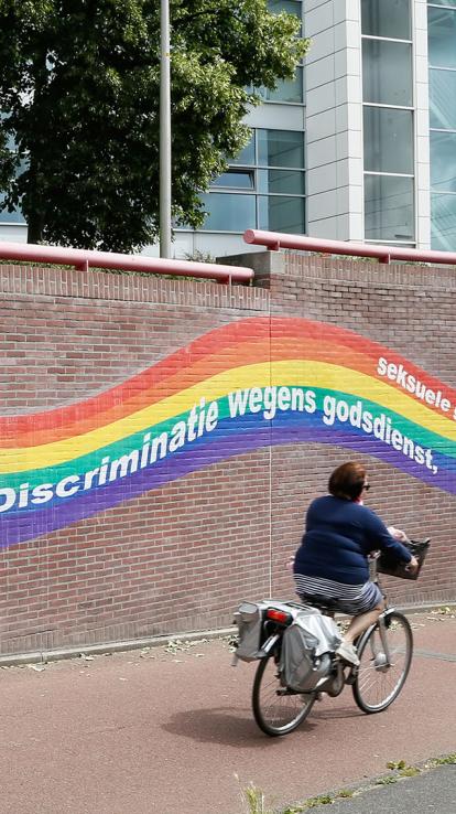 Vrouw op fiets voor een muur met de regenboogvlag waar teksten opstaan over discriminatie.  