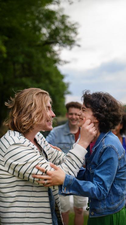 Twee vrienden zijn samen. De een houdt de armen van de ander vast, terwijl de ander zachtjes in de wangen van zijn vriend knijpt.