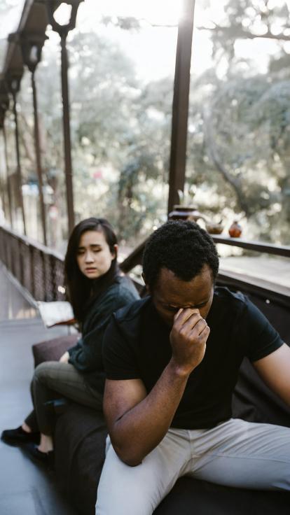 Twee vrienden zitten op een bank zichtbaar van streek. De man op de voorgrond houdt zijn hoofd in zijn hand en lijkt gefrustreerd. De vrouw achter hem kijkt bezorgd naar hem, alsof ze midden in een moeilijke discussie zitten.