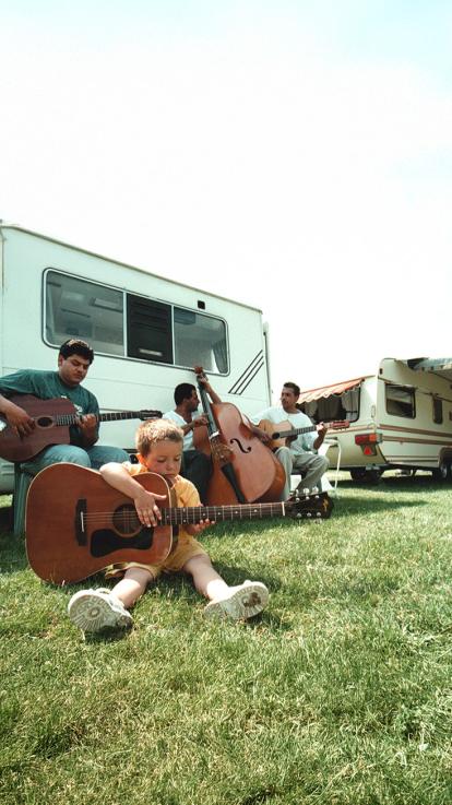 Een groep mensen speelt gitaar en contrabas naast woonwagens. Een jong kind probeert ook een gitaar te bespelen op het gras.
