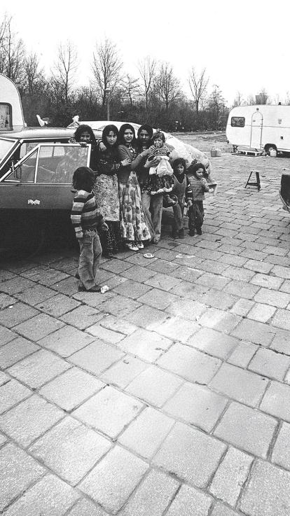 Een Sinti-familie poseert samen bij een auto en woonwagens. Kinderen en volwassenen staan dicht bij elkaar op een geplaveide ondergrond.