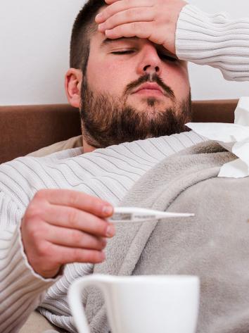 een zieke man in bed, met hand op voorhoofd en thermometer in andere hand