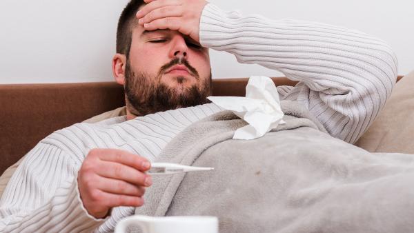 een zieke man in bed, met hand op voorhoofd en thermometer in andere hand