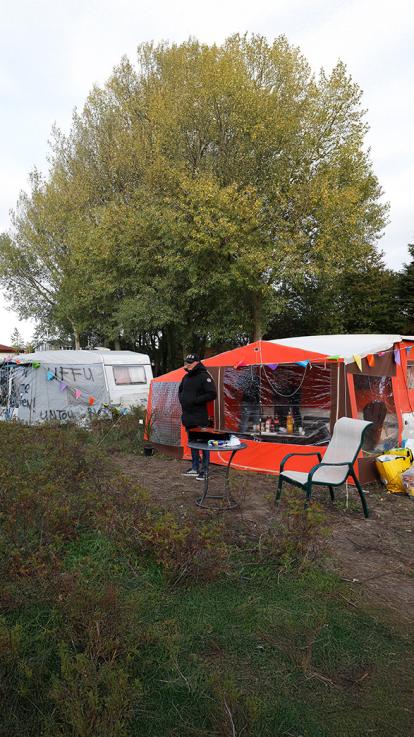 Een woonwagenkamp met een man naast een oranje voortent, vlaggetjes en protestborden. De woonwagens staan tussen bomen en struiken.