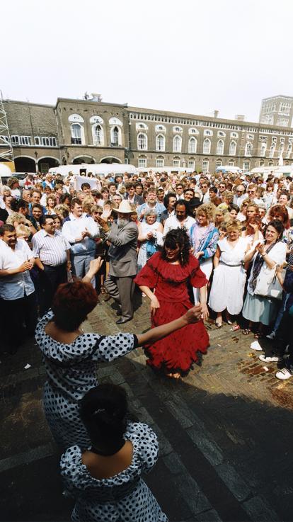 Vrouw in rode jurk danst op een plein, omringd door een grote menigte mensen. Andere dansers in traditionele kleding kijken toe.