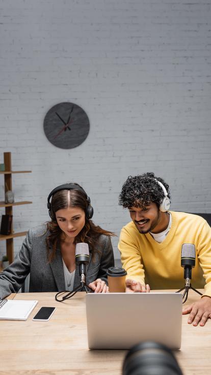 Twee mensen met koptelefoon op achter microfoons en een laptop nemen een podcast op