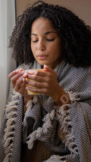 Vrouw met kleedje om haar heen en kop thee zit tegen de verwarming aan