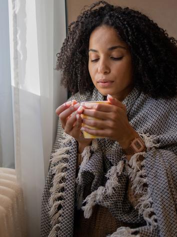 Vrouw met kleedje om haar heen en kop thee zit tegen de verwarming aan