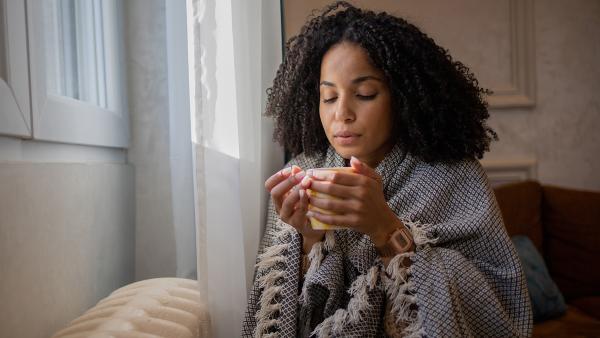 Vrouw met kleedje om haar heen en kop thee zit tegen de verwarming aan