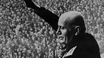 een zwart-wit foto van Mussolini die zijn hand opsteekt richting een grote mensenmassa vanaf een balkon