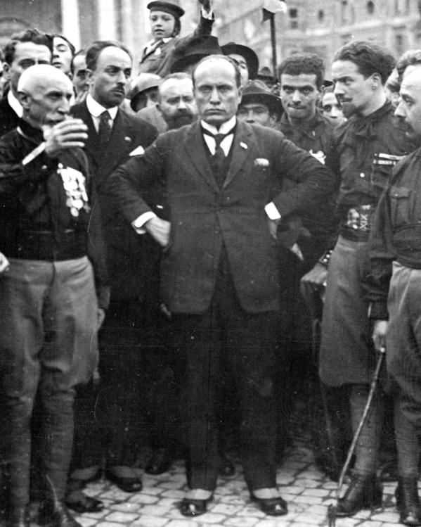 Mussolini met zijn handen in zijn zij, middenin een groep aanhangers